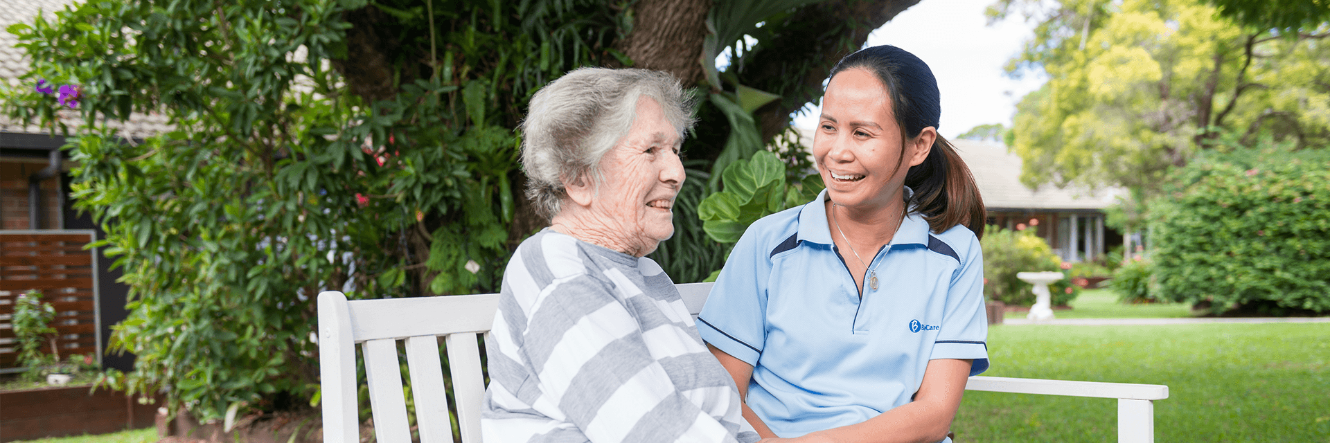 A smiling Blue Care employee holding hands with an older woman on a park bench