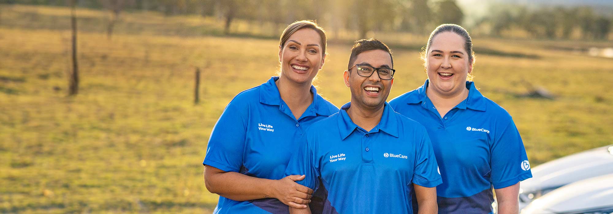 Three BlueCare staff standing together in a field in front of a fleet of BlueCare vehicles, smiling at camera